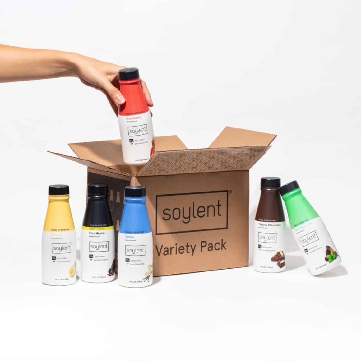 A hand placing a bottle of Soylent plant-based ready-to-drink shake into a cardboard box with five other shake flavors next to the box.