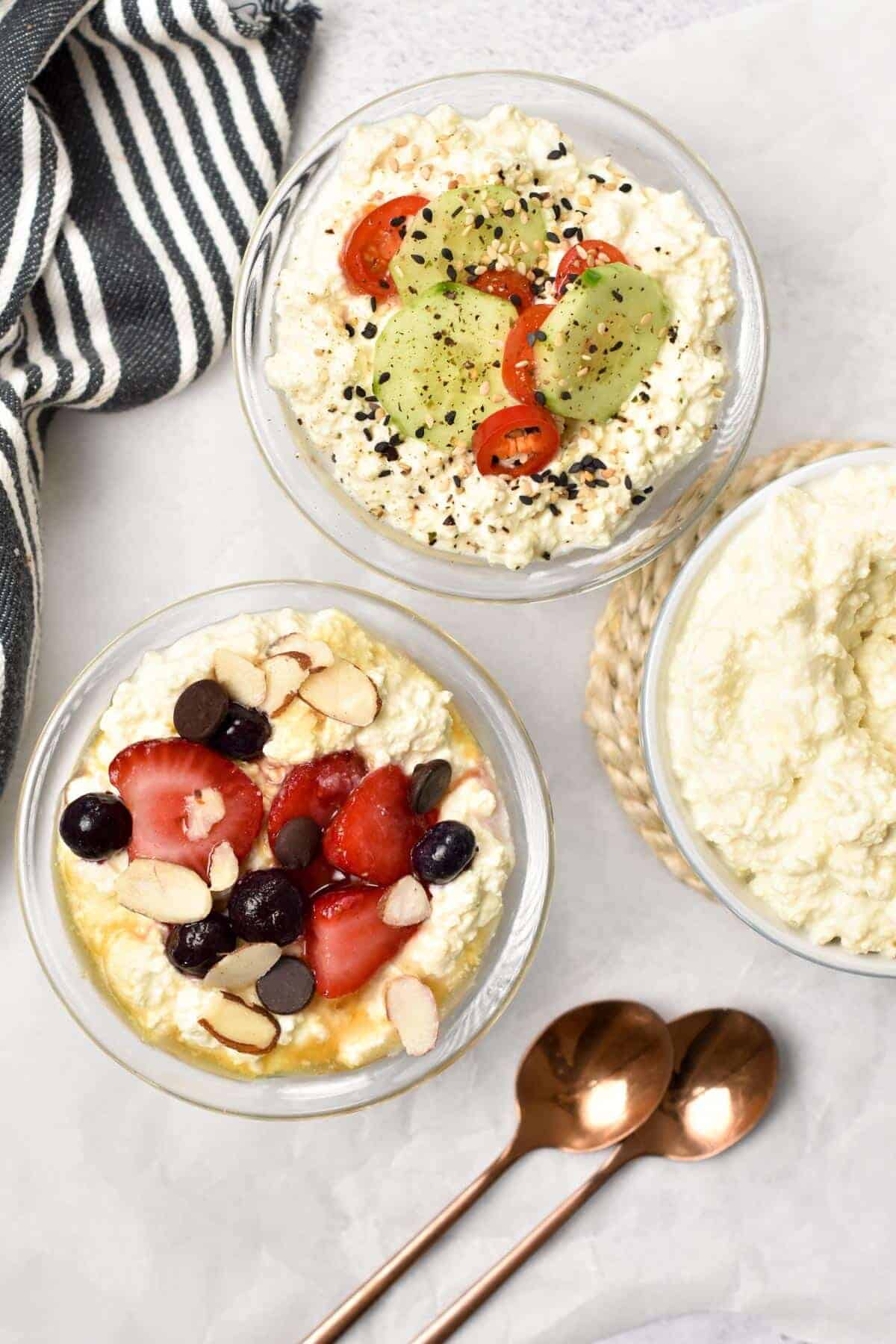 Three glass bowls filled with vegan cottage cheese and various fruit and vegetable toppings on a white background and accented by copper spoons and a gray and white striped towel.