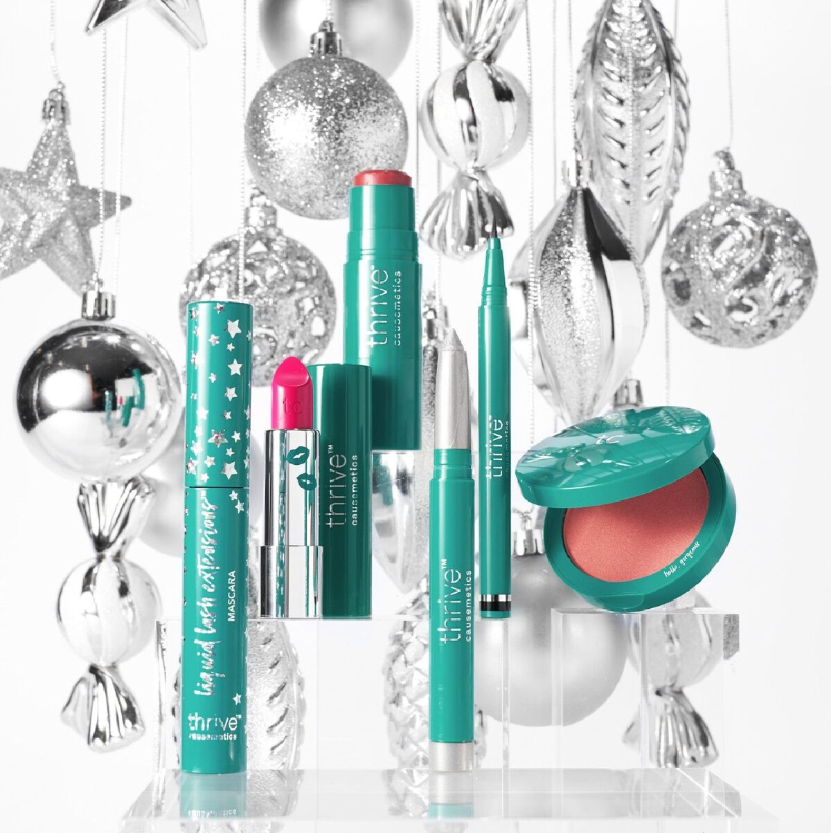 Green Thrive Causemetics Liquid Lash Extender Mascara next to other Thrive Causemetic beauty products against a white background with silver hanging ornaments. 