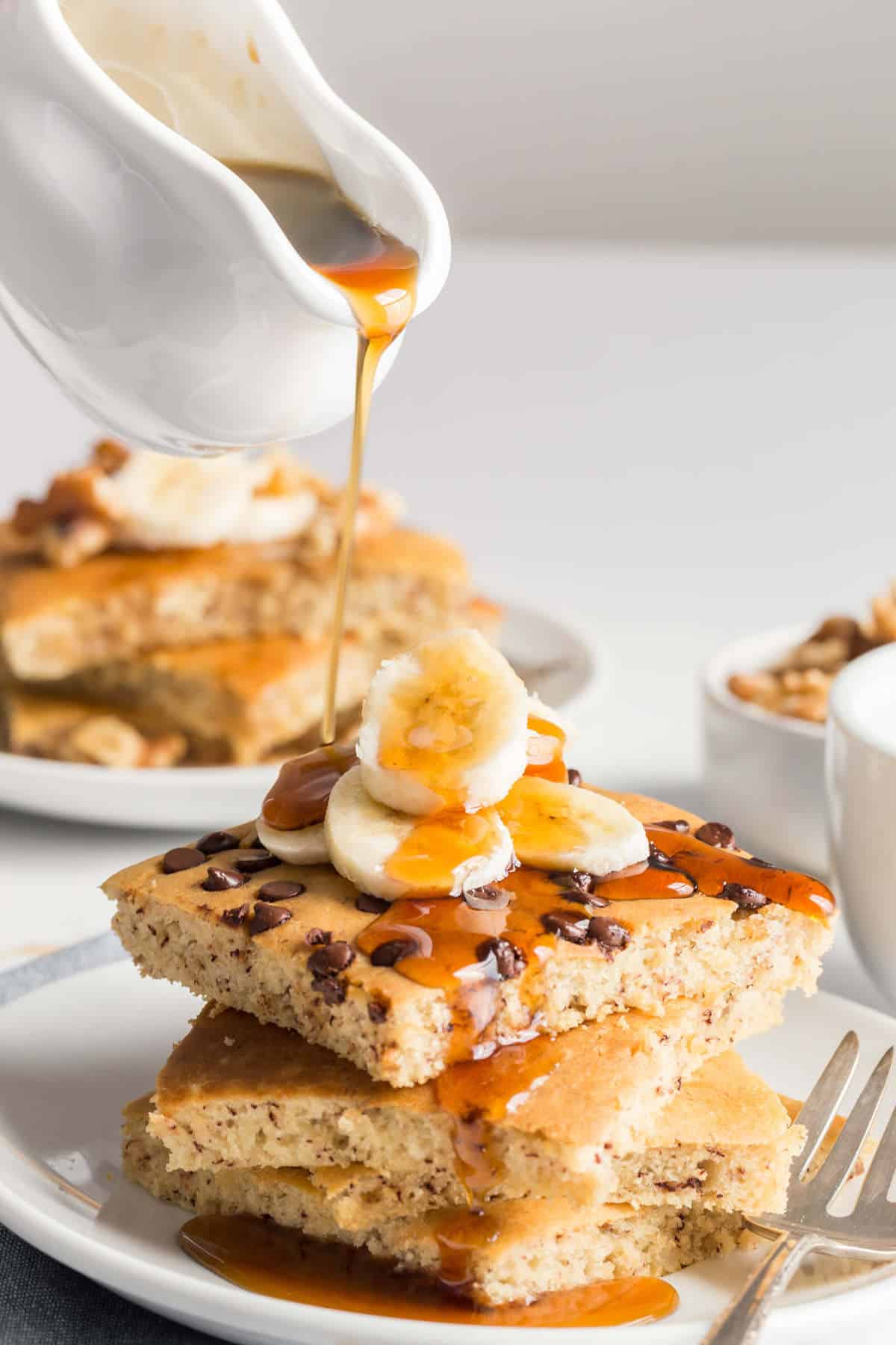 Vegan sheet pan pancakes stacked on a plate, topped with banana slices and maple syrup.