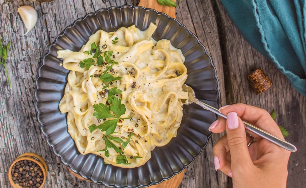 A plate of vegan fettuccine alfredo with a hand holding a fork.