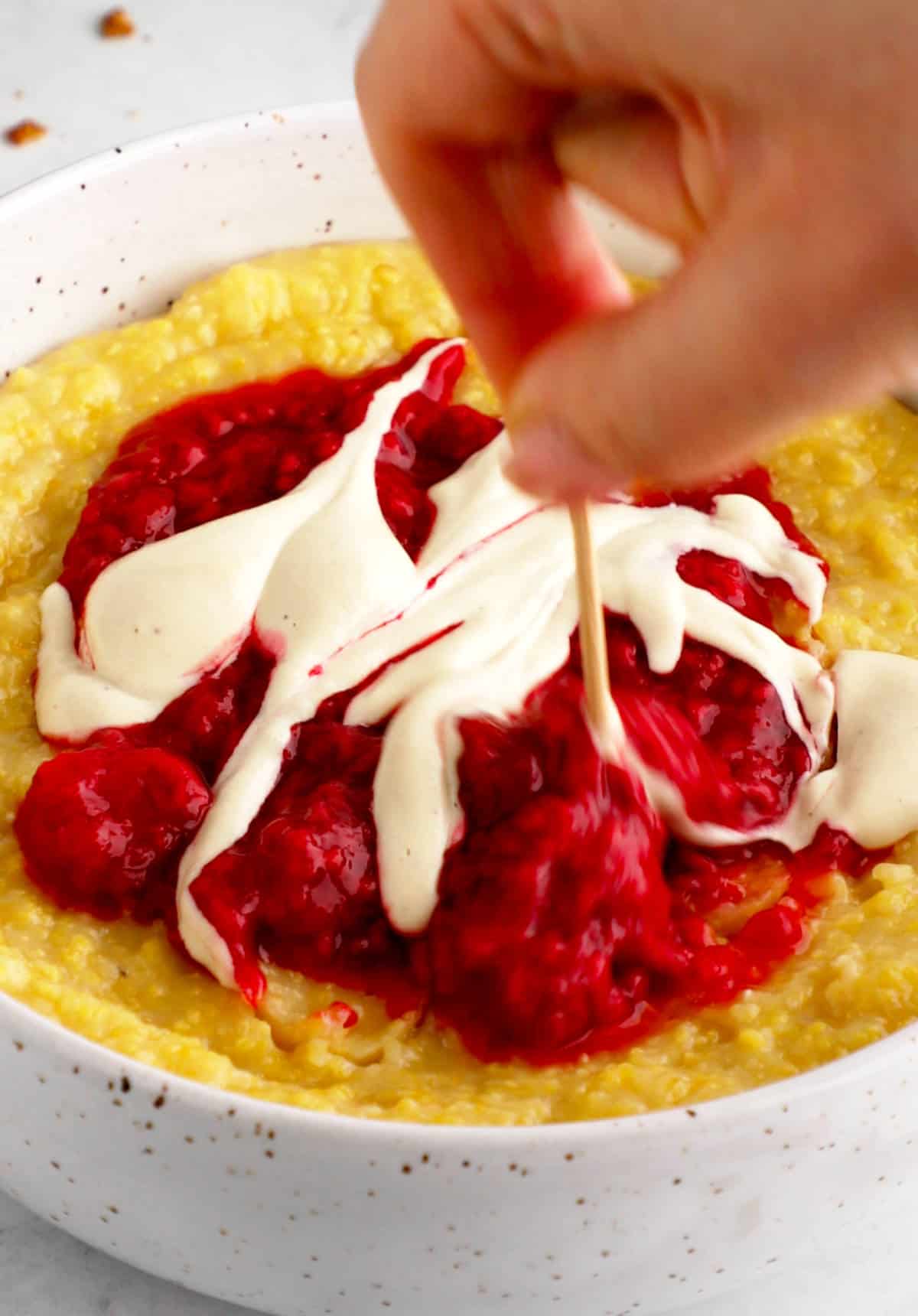 Cashew cream drizzled over the berry compote vegan grits bowl with a hand using a toothpick to swirl it around.