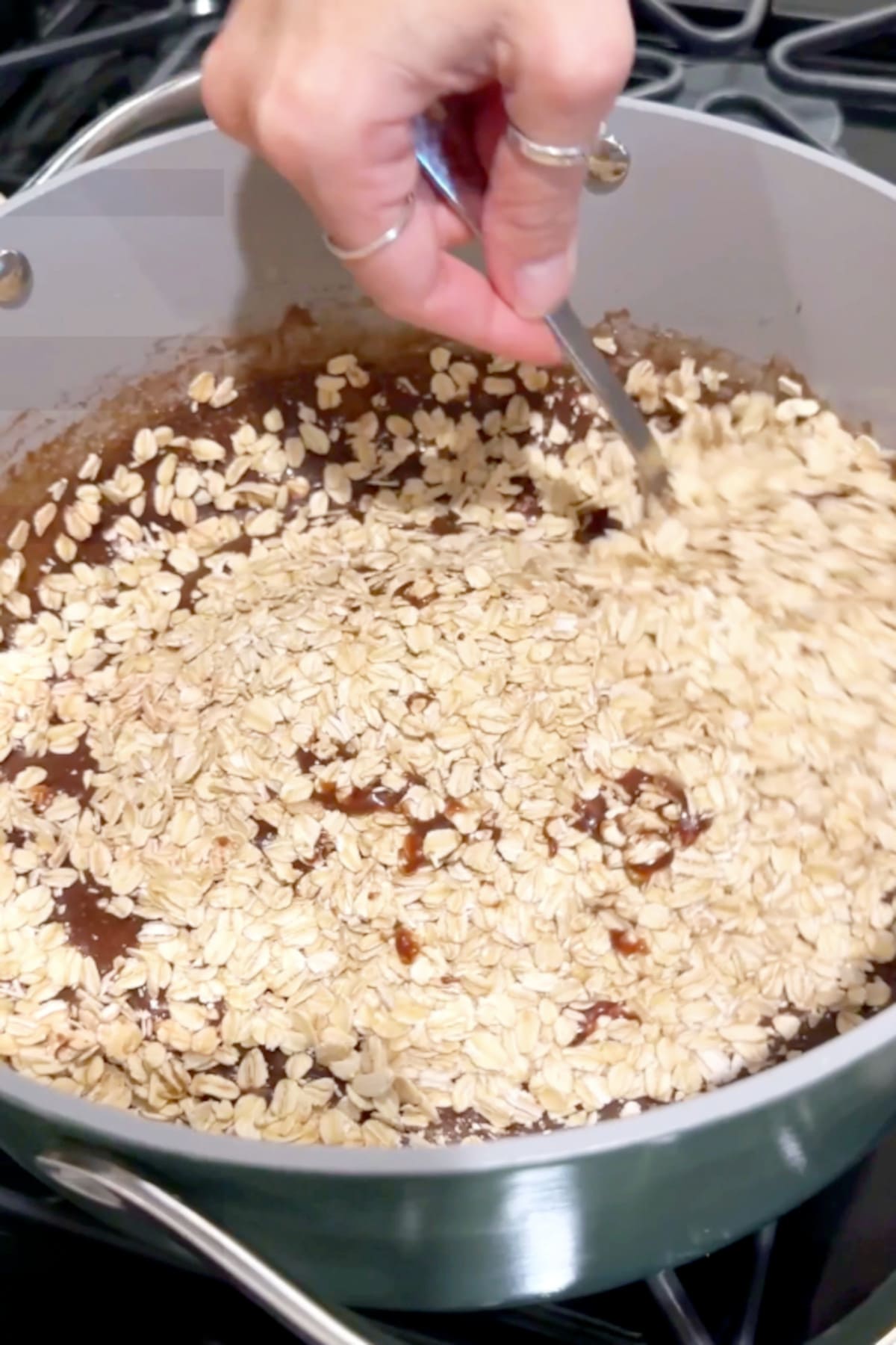 A hand using a spoon to stir the rolled oats into the cacao mixture in the pan.
