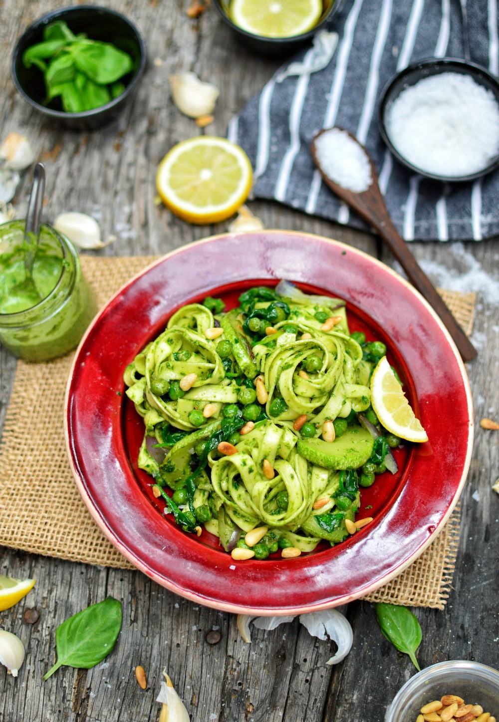 A bowl of vegan pesto pasta served on a cutting board surrounded by various pesto ingredients.