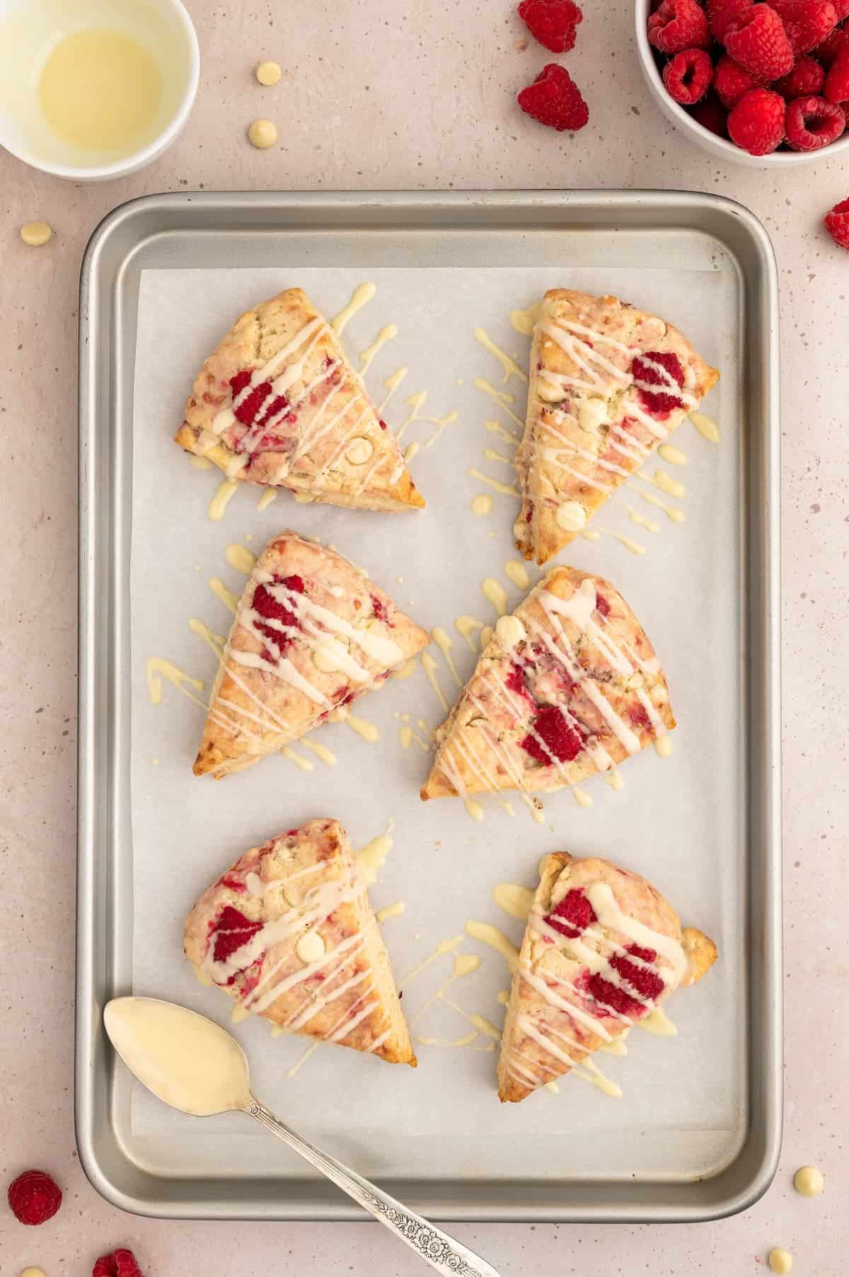 Raspberry vegan scones on a baking sheet drizzled with melted vegan white chocolate.