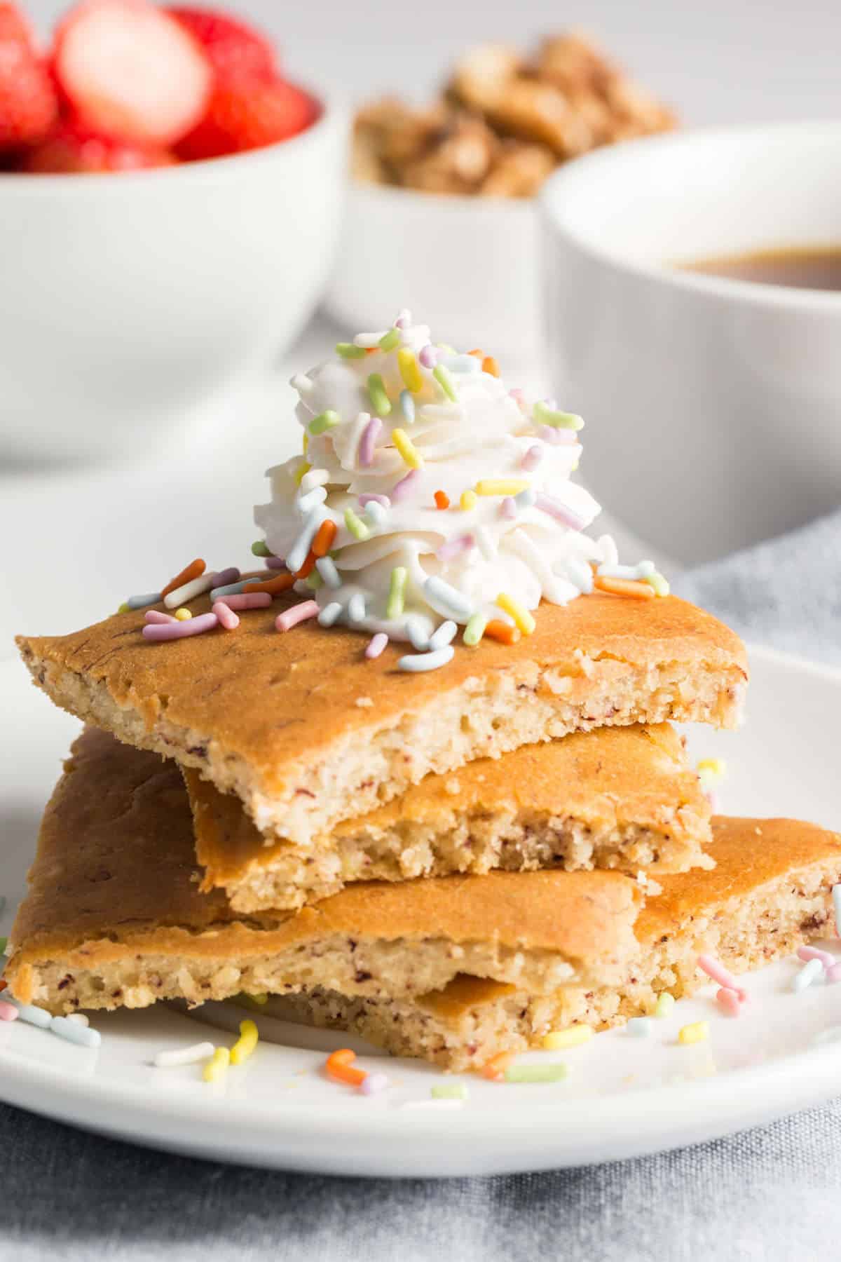 A stack of vegan sheet pan pancakes on a plate topped with sprinkles and vegan whipped cream.