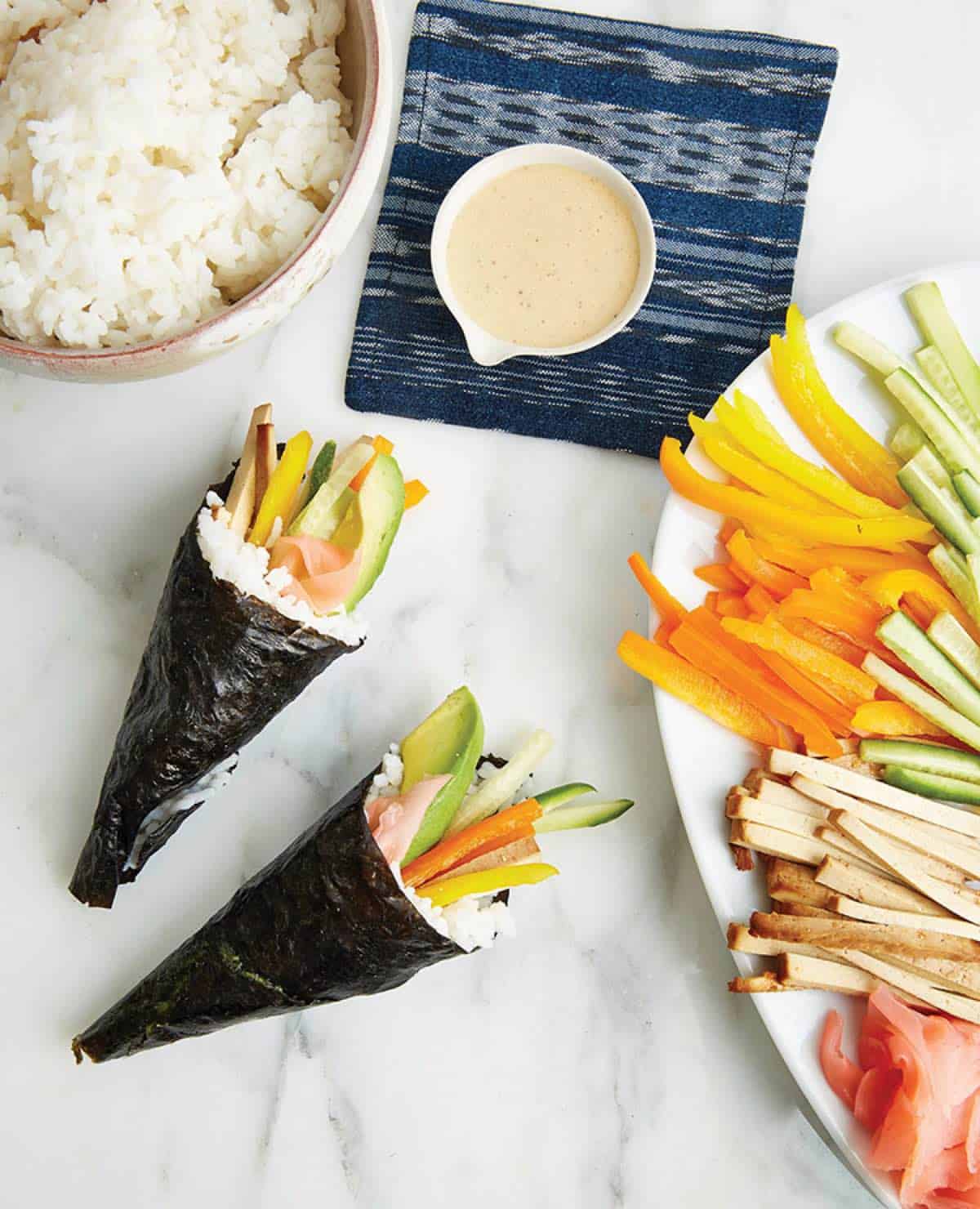 Vegan sushi hand rolls with baked tofu and veggies on a table with rice.