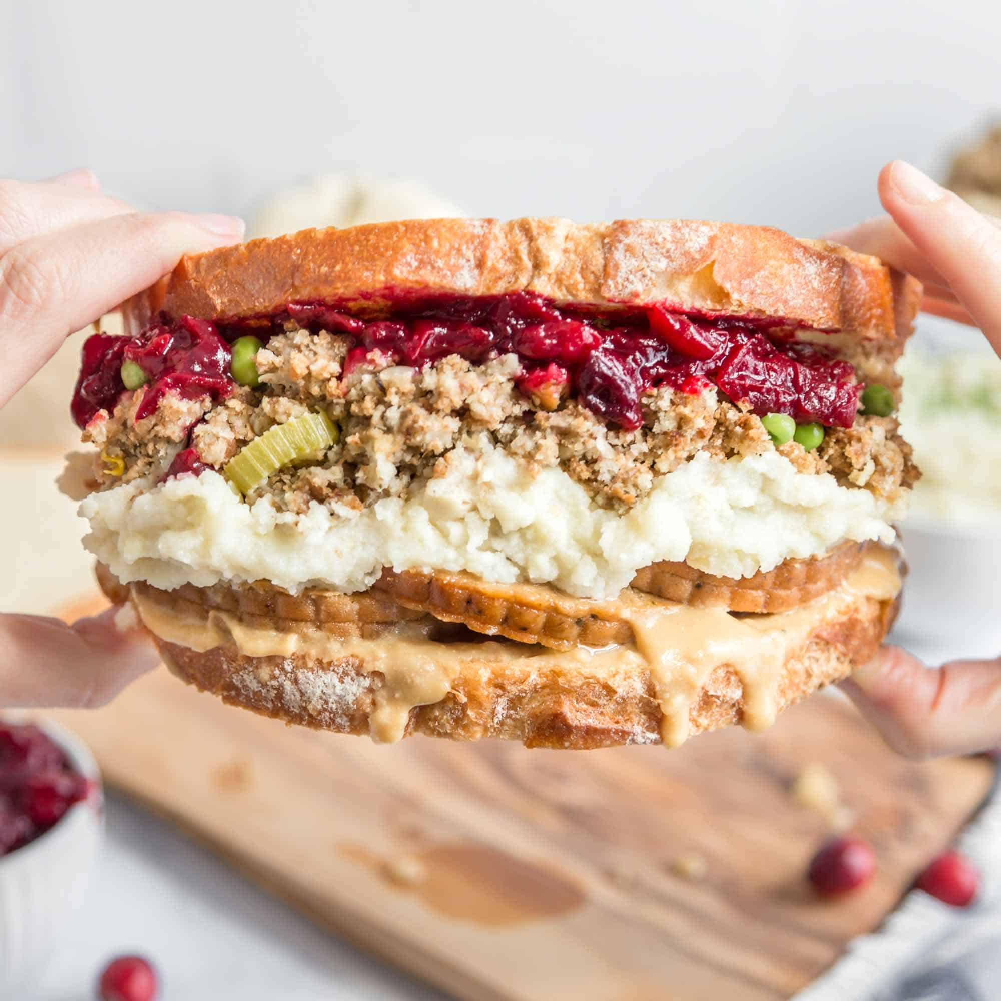 hands holding a thanksgiving leftovers sandwich stuffed with mashed potatoes, stuffing, vegan turkey, and cranberry sauce