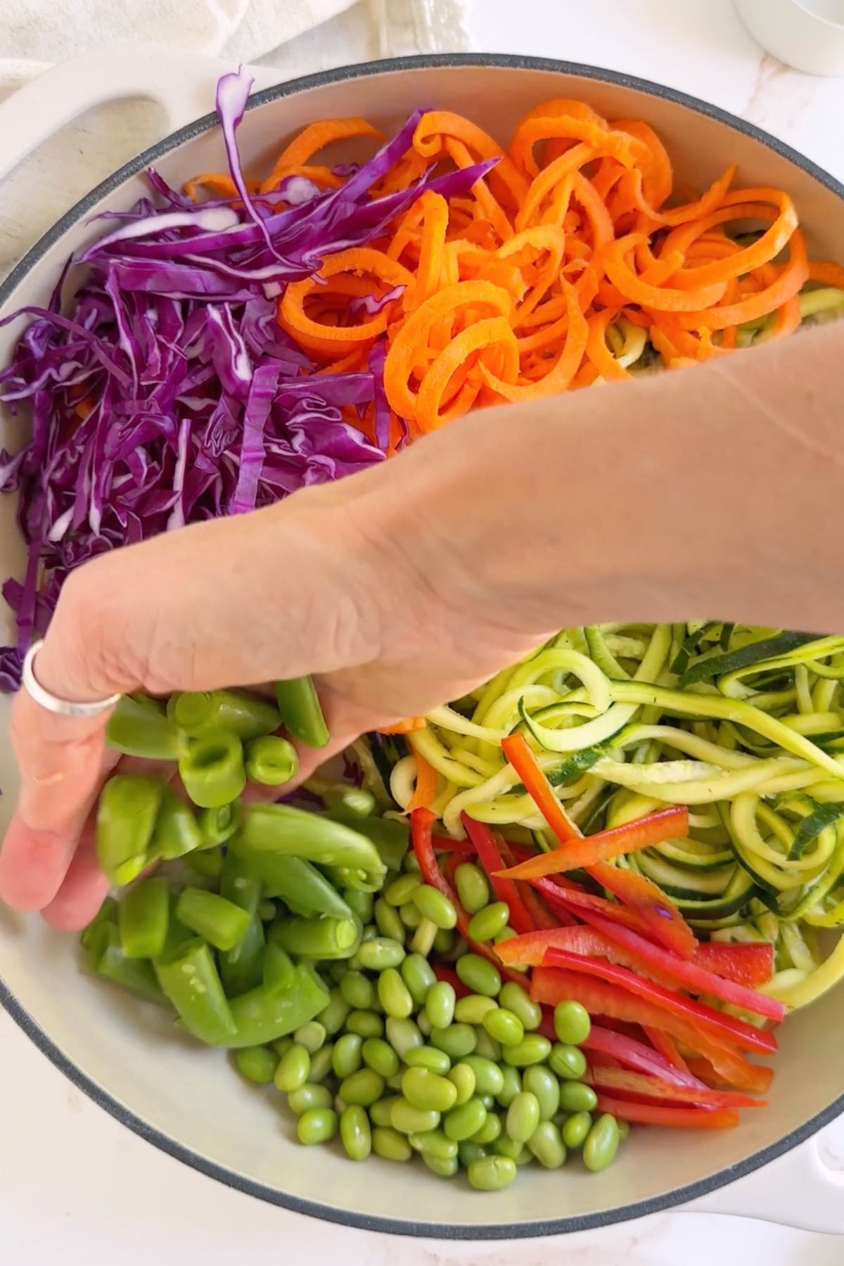 A rainbow bed of fresh veggies including spiralized zoodles, carrot noodles, red bell pepper, edamame, snap peas, and purple cabbage. 
