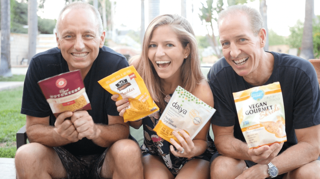 Greg Hicks, Michelle Cehn, and Dan Safko hold up four bags of vegan shredded cheddar cheese